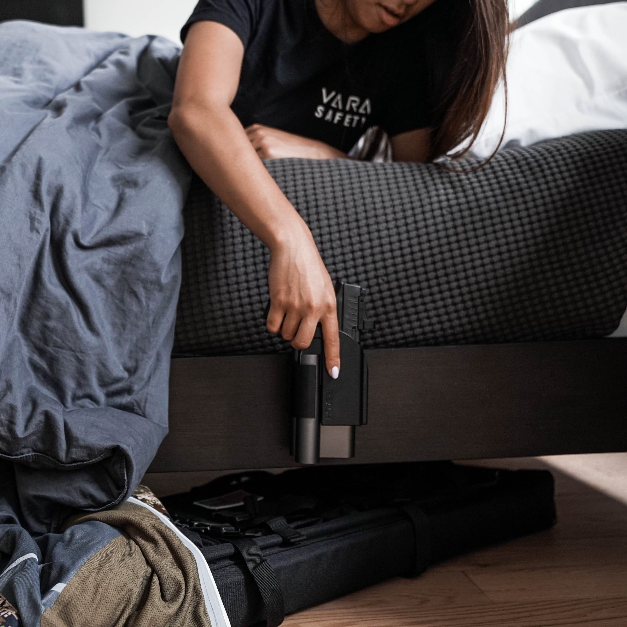 A woman reaching down from bed to a Reach 2S handgun safe mounted on the bed frame